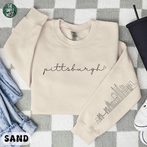 Op de afbeelding: Een zandkleurige sweatshirt met het woord "Pittsburgh" in cursief schrift. De mouw heeft een zwarte lijntekening van de skyline van Pittsburgh. Het logo The Simple West staat in de linkerbovenhoek.