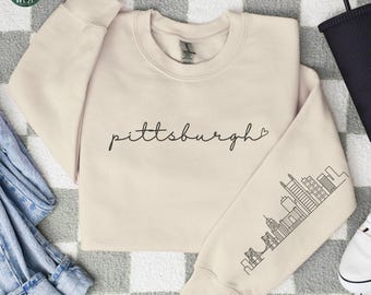 May include: A sand-colored sweatshirt with the word "Pittsburgh" in cursive script. The sleeve features a black line drawing of the Pittsburgh skyline. The Simple West logo is in the upper left corner.