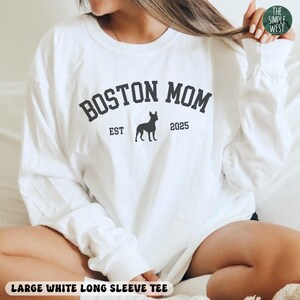 Puede incluir: Camiseta blanca de manga larga con el texto "BOSTON MOM" en gris oscuro, una silueta de perro y el a&ntilde;o "2025". La camiseta tambi&eacute;n tiene el texto "LARGE WHITE LONG SLEEVE TEE" en la parte inferior.