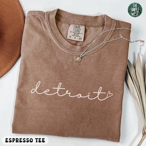 May include: Espresso-colored t-shirt with the word "Detroit" in cursive script. The shirt has a crew neck and a small tag at the neckline. A silver necklace with a pendant is draped across the shirt. The shirt is labeled "Espresso Tee".