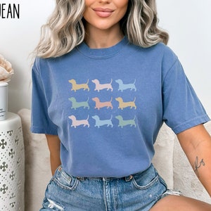 May include: A blue jean t-shirt featuring a design of nine pastel-coloured dachshund silhouettes. The t-shirt is being worn by a person with long blonde hair.
