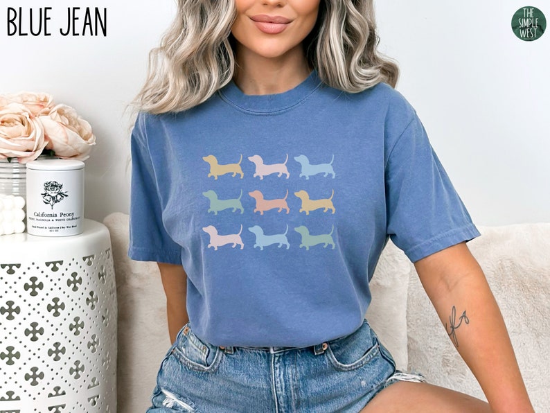 May include: A blue jean t-shirt featuring a design of nine pastel-coloured dachshund silhouettes. The t-shirt is being worn by a person with long blonde hair.