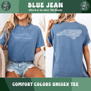 Pu&ograve; includere: T-shirt unisex Comfort Colors blu jeans. La parte anteriore della maglietta blu ha le parole "north carolina" in corsivo bianco. Il retro della maglietta presenta un contorno bianco dello stato della Carolina del Nord con un design decorativo. Il logo The Simple West &egrave; negli angoli superiori.