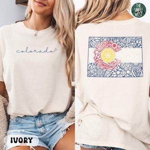 May include: Ivory-colored t-shirt with the word "colorado" in blue script on the front. The back features a Colorado flag design with floral patterns in blue, red, and yellow. The Simple West logo is in the upper right corner.