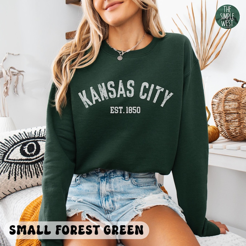 K&ouml;nnte beinhalten: Ein dunkelgr&uuml;ner Pullover mit dem Text "Kansas City Est. 1850" in Wei&szlig;. Der Pullover wird von einer Person mit einem hellblauen Jeansrock getragen.