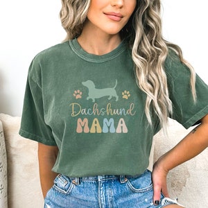 Puede incluir: Una camiseta verde con una silueta de dachshund y el texto "Dachshund Mama" en letras de colores.