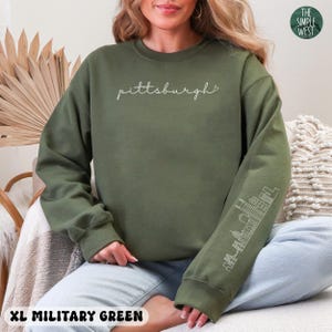 Op de afbeelding: Militairgroen sweatshirt met het woord "Pittsburgh" in cursief schrift. De mouw heeft een witte omtrek van de skyline van de stad. De tekst "XL MILITARY GREEN" staat onderaan de afbeelding. Het logo The Simple West staat rechtsboven.