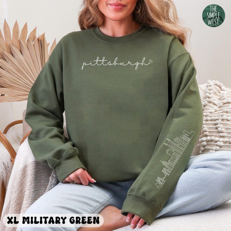 Op de afbeelding: Militairgroen sweatshirt met het woord "Pittsburgh" in cursief schrift. De mouw heeft een witte omtrek van de skyline van de stad. De tekst "XL MILITARY GREEN" staat onderaan de afbeelding. Het logo The Simple West staat rechtsboven.