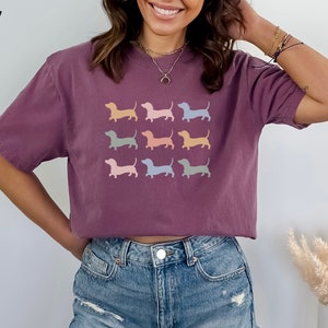 May include: A purple cropped t-shirt featuring a graphic of nine pastel coloured dachshund silhouettes. The text "BERRY" is in the top left corner of the image.