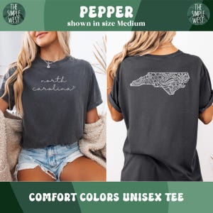 May include: Pepper-colored Comfort Colors unisex tee. The front features the text "north carolina" in a cursive font. The back displays a white, detailed outline of North Carolina. The shirt is shown in size Medium.