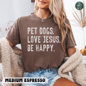 Puede incluir: Camiseta de color espresso medio con el texto "PET DOGS. LOVE JESUS. BE HAPPY." en letras blancas. La camiseta es de cuello redondo y tiene un corte relajado. El logotipo de The Simple West est&aacute; en la esquina superior derecha.