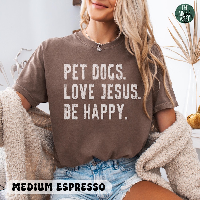 Puede incluir: Camiseta de color espresso medio con el texto "PET DOGS. LOVE JESUS. BE HAPPY." en letras blancas. La camiseta es de cuello redondo y tiene un corte relajado. El logotipo de The Simple West est&aacute; en la esquina superior derecha.