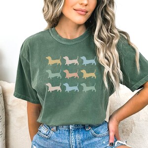 May include: A green t-shirt featuring a graphic of several dachshund silhouettes in a range of pastel colours. The text "MOSS" is in the top left corner and "THE SIMPLE WEST" is in the top right corner.
