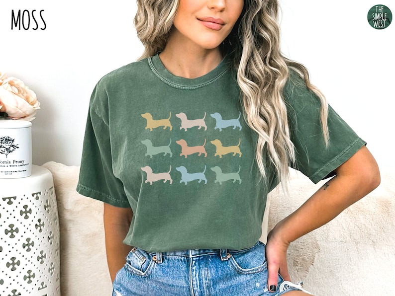 May include: A green t-shirt featuring a graphic of several dachshund silhouettes in a range of pastel colours. The text "MOSS" is in the top left corner and "THE SIMPLE WEST" is in the top right corner.