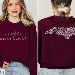 May include: Maroon sweatshirt with the text "north carolina" in white script on the front and a white outline of the state of North Carolina with a floral design on the back.