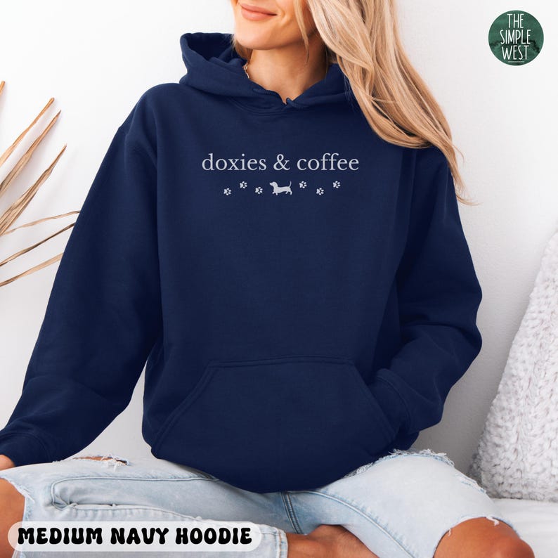 May include: Medium navy blue hoodie with a front pocket and a drawstring hood. The hoodie features the text "doxies & coffee" in white, along with paw prints and a dachshund silhouette. The Simple West logo is in the upper right corner.