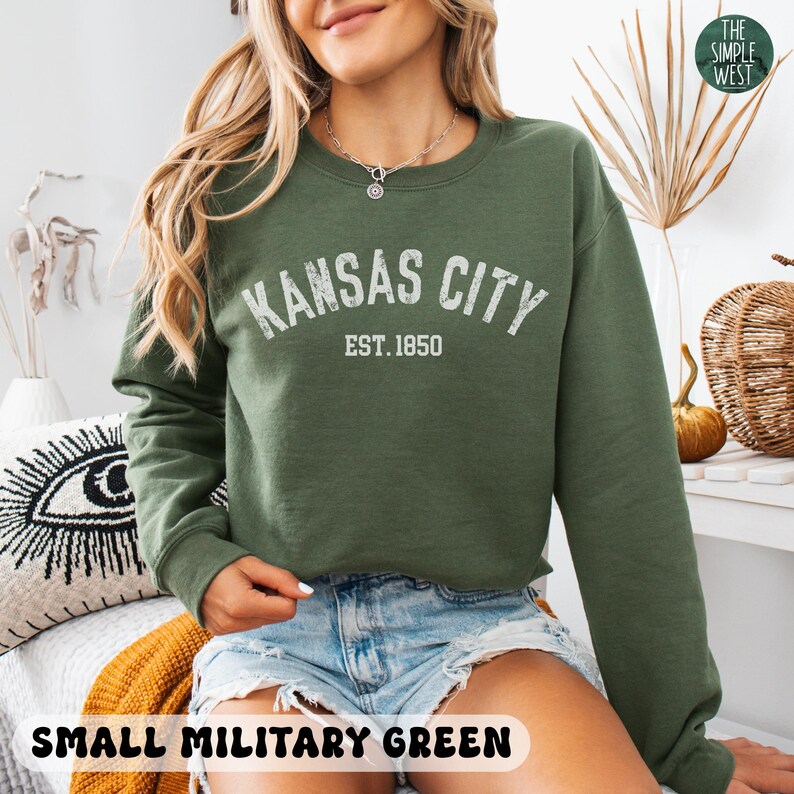K&ouml;nnte beinhalten: Ein gr&uuml;ner Pullover mit dem Text "Kansas City Est. 1850" in Wei&szlig;. Das Model tr&auml;gt den Pullover mit einer Jeans-Shorts.