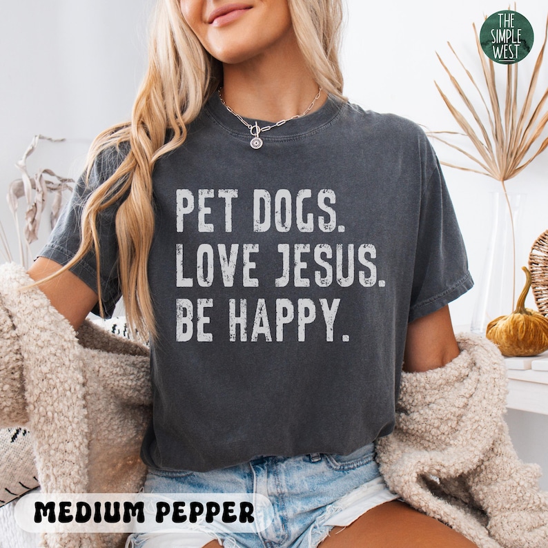 Puede incluir: Camiseta gris oscuro con el texto "PET DOGS. LOVE JESUS. BE HAPPY." en blanco. La camiseta es de color pimienta media. El logotipo de The Simple West est&aacute; en la esquina superior derecha. La persona lleva un collar y un su&eacute;ter beige.