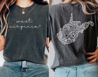 Puede incluir: Una camiseta gris oscuro con las palabras "west virginia" en escritura blanca en la parte delantera y un contorno blanco del estado de Virginia Occidental con un diseño floral en la parte posterior. La camiseta se muestra en una modelo que lleva pantalones vaqueros azules.