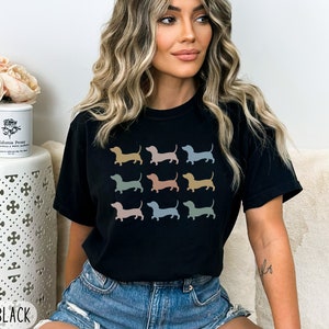 May include: Black t-shirt featuring a graphic print of several dachshund dogs in a variety of colours. The text "MEDIUM BLACK" is visible at the bottom of the image.