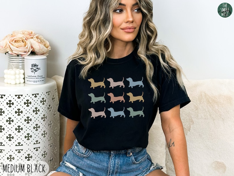 May include: Black t-shirt featuring a graphic print of several dachshund dogs in a variety of colours. The text "MEDIUM BLACK" is visible at the bottom of the image.