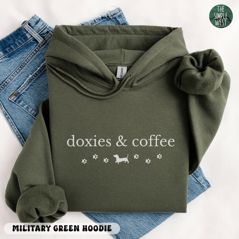 May include: Military green hoodie with the text "doxies & coffee" and a dachshund illustration. The hoodie has a drawstring hood and long sleeves. The Simple West logo is in the upper right corner.