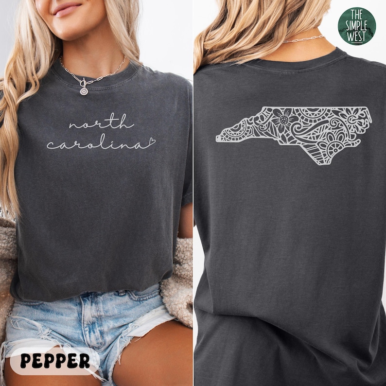 Pu&ograve; includere: T-shirt color pepe con le parole "north carolina" in corsivo. Il retro della maglietta presenta un contorno bianco e dettagliato della forma dello stato della Carolina del Nord. Il logo The Simple West &egrave; visibile.
