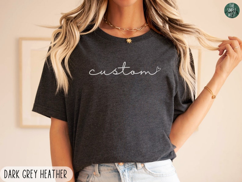以下が含まれることがあります： 「custom」の文字が白い筆記体で書かれた、ダークグレーのヘザーTシャツ。The Simple Westのロゴが右上にある。このシャツはクルーネックです。