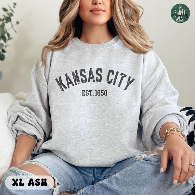 K&ouml;nnte beinhalten: Ein graues Sweatshirt mit dem Text "KANSAS CITY EST. 1850" in Schwarz. Das Sweatshirt wird von einer Person getragen.