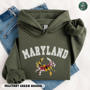 May include: Military green hoodie with the word "MARYLAND" in white block letters and a crab graphic in the center. The crab graphic features the Maryland state flag colors. The hoodie is folded on top of blue jeans.