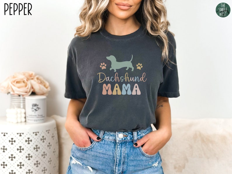 Puede incluir: Una camiseta gris oscuro con una silueta de dachshund y huellas de patas en colores pastel. El texto "Dachshund Mama" est&aacute; impreso en una fuente retro colorida.