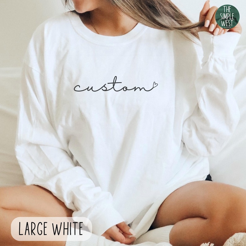 May include: A white long-sleeve shirt with the word "custom" in a cursive font. The shirt has a small heart at the end of the word. The text "LARGE WHITE" is at the bottom of the image. The Simple West logo is in the top right corner.