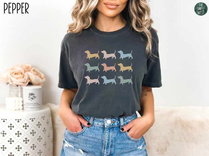 May include: A dark grey t-shirt featuring a graphic of several pastel-coloured dachshund silhouettes. The text "PEPPER" is in the top left corner of the image.
