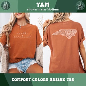Pu&ograve; includere: T-shirt unisex Comfort Colors arancione bruciato. La parte anteriore della maglietta presenta la scritta "north carolina" in corsivo. Il retro della maglietta presenta un contorno bianco e dettagliato della Carolina del Nord. Il logo The Simple West si trova negli angoli superiori.