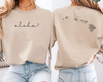 Könnte beinhalten: Ein mittel sandfarbenes Sweatshirt mit dem Wort "aloha" in Schreibschrift auf der Vorderseite. Die Rückseite zeigt eine schwarze Umrisszeichnung der hawaiianischen Inseln. Das Logo The Simple West ist sichtbar. Das Sweatshirt wird mit hellblauen Jeans kombiniert.