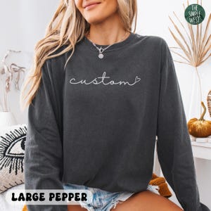 May include: Dark gray sweatshirt with the word "custom" in white script. The sweatshirt has a crew neck and long sleeves. The text "LARGE PEPPER" is at the bottom of the image. The Simple West logo is in the top right corner.