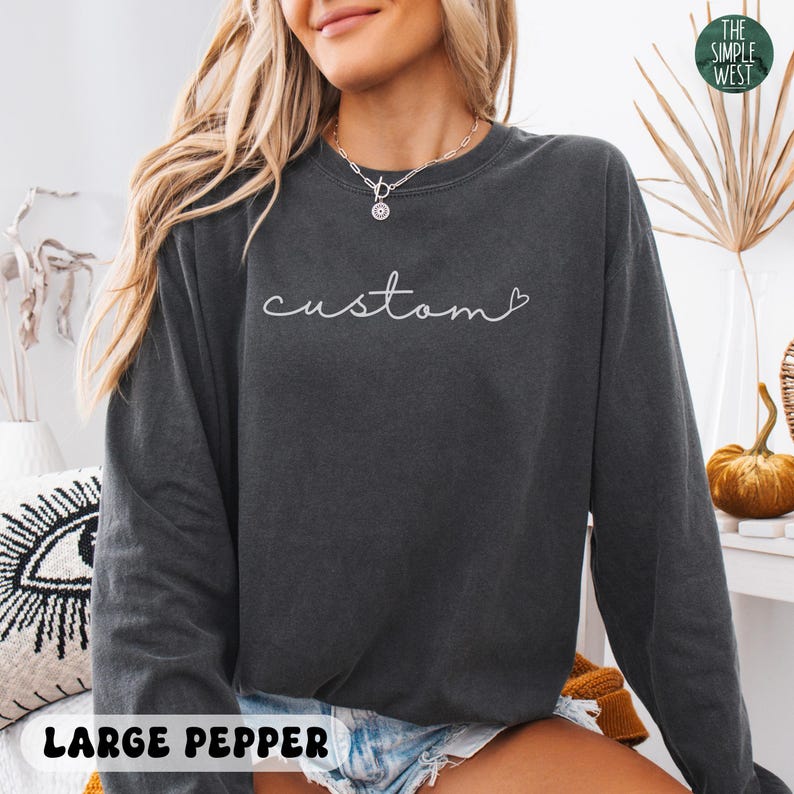 May include: Dark gray sweatshirt with the word "custom" in white script. The sweatshirt has a crew neck and long sleeves. The text "LARGE PEPPER" is at the bottom of the image. The Simple West logo is in the top right corner.