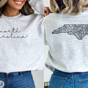 May include: A grey sweatshirt with the text "north carolina" on the front and a black and white outline of the state of North Carolina with a floral design on the back. The sweatshirt is shown on a model wearing blue jeans.