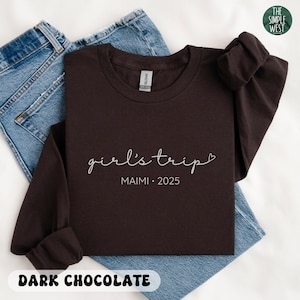 May include: A dark chocolate-colored long-sleeve shirt with the words "girl's trip" and "MAIMI &bull; 2025" printed in white script. The shirt is folded on top of a pair of blue jeans. The words "DARK CHOCOLATE" are printed at the bottom.