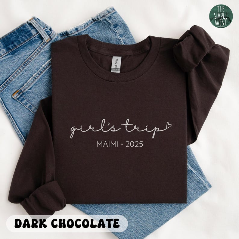 May include: A dark chocolate-colored long-sleeve shirt with the words "girl's trip" and "MAIMI &bull; 2025" printed in white script. The shirt is folded on top of a pair of blue jeans. The words "DARK CHOCOLATE" are printed at the bottom.
