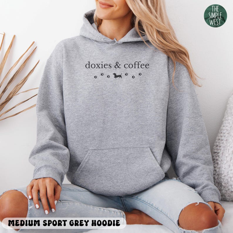 May include: Medium sport grey hoodie with a front pocket and a drawstring hood. The hoodie features the text "doxies & coffee" above a dachshund illustration and paw prints. The Simple West logo is in the upper right corner.