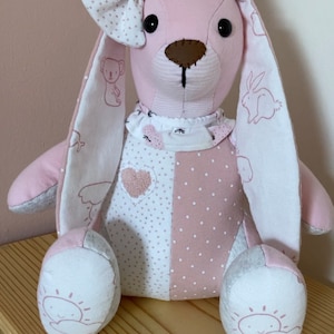 Könnte beinhalten: Ein rosa-weißes Patchwork-Plüschkaninchen mit einer weißen Schleife. Das Kaninchen hat gestickte Details an den Ohren und Füßen, darunter eine Sonne, Wolken und ein Herz.