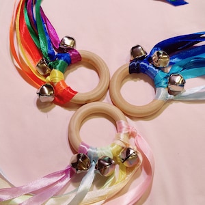 May include: Three wooden rings with colorful ribbons and bells attached. The rings are made of natural wood and have a smooth finish. The ribbons are tied to the rings and have bells at the ends. The ribbons are in a variety of colors, including red, orange, yellow, green, blue, indigo, and violet. The bells are silver and have a jingle sound.