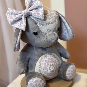 May include: A gray plush elephant toy with a large bow on its head. The elephant has floral patterned fabric on its ears, head, belly, and feet. The bow and patterned fabric feature a white and light purple floral design. The elephant is sitting on a wooden base.