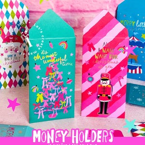 Op de afbeelding: Een verzameling kleurrijke geldhouders met kerstthema's. Eén teal houder heeft een roze kerstboomillustratie en de tekst "It's the most wonderful time". Een andere roze houder heeft rode strepen en een notenkrakerillustratie met de tekst "All I want for Christmas is you".