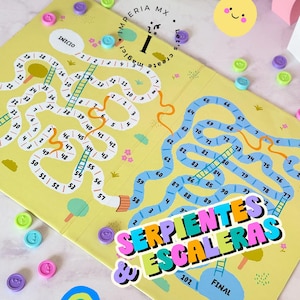Puede incluir: Un juego de mesa amarillo con un camino sinuoso, escaleras y números. El tablero del juego presenta el texto "SERPIENTES & ESCALERAS" y está rodeado de coloridas fichas y una pegatina de arcoíris. El juego es para varios jugadores.