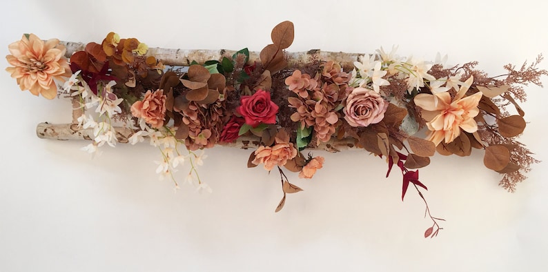 May include: A rustic wooden ladder decorated with a variety of artificial flowers in shades of peach, pink, red, and brown. The flowers are arranged in a cascading style, creating a beautiful and natural look.