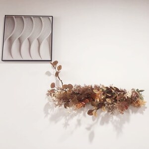 May include: A white wall with a framed piece of art featuring a wavy pattern and a garland of dried flowers and leaves.