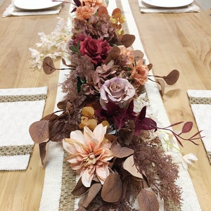 May include: A floral centerpiece with a mix of pink, red, and orange flowers, brown leaves, and white sprigs. The centerpiece is on a white and brown table runner.