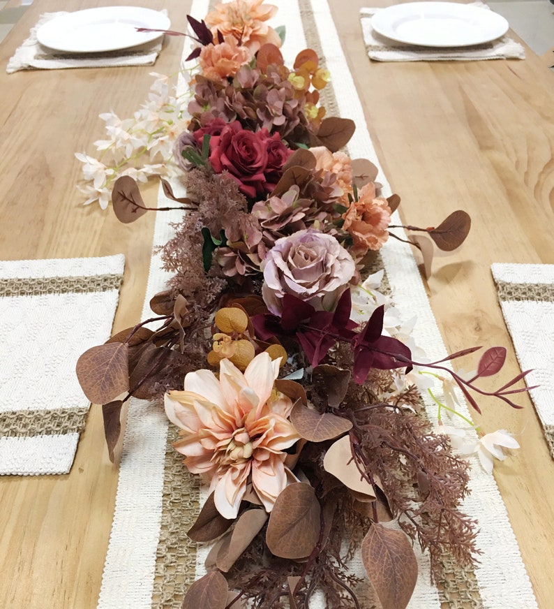 May include: A floral centerpiece with a mix of pink, red, and orange flowers, brown leaves, and white sprigs. The centerpiece is on a white and brown table runner.